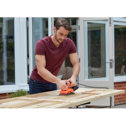 Muž brousí dřevěné dveře s vibrační bruskou Black+Decker BEW220 150 W. Bruska na dřevo.